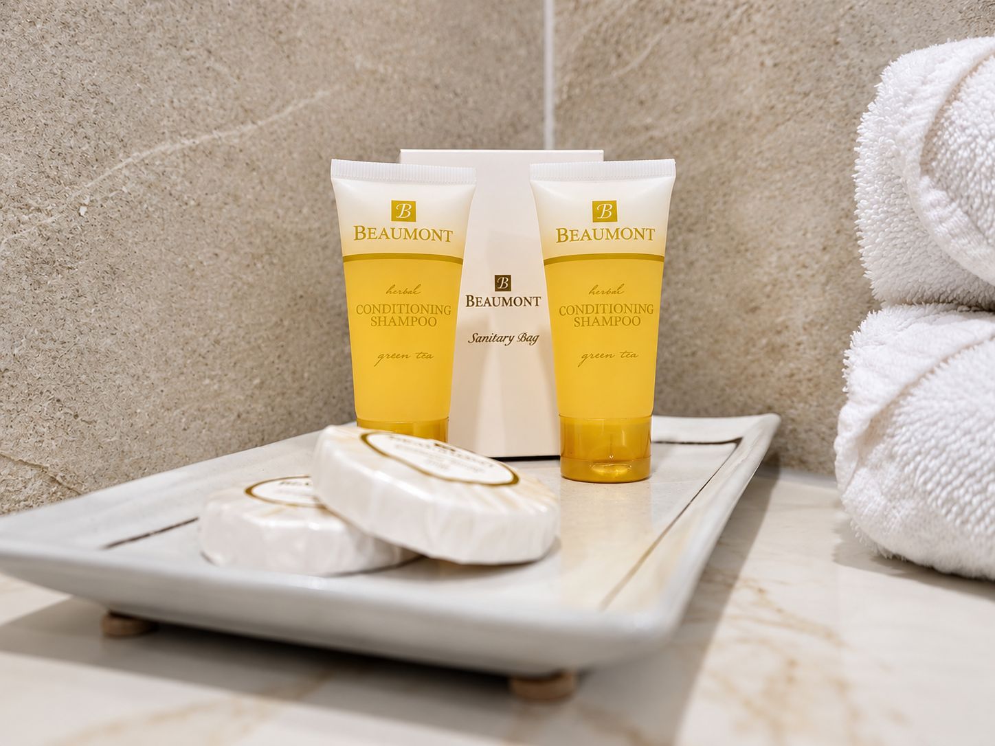 A bathroom counter with a white ceramic tray holding a bar of soap and two tubes of Beaumont conditioning shampoo.