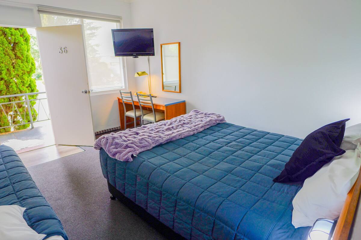 A bedroom with a bed, a desk, a chair, a TV, and a door.