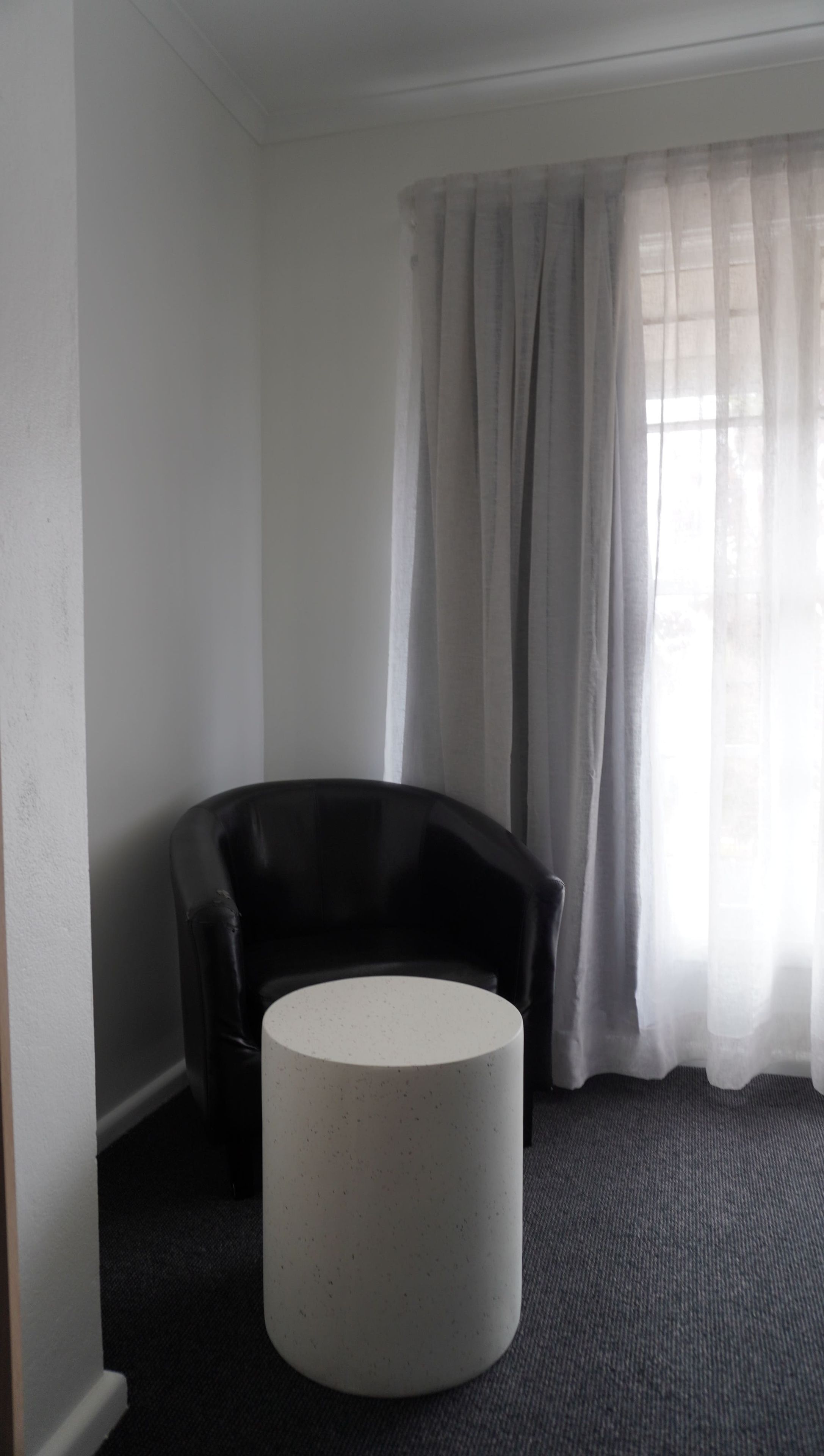 Ein Zimmer mit einem schwarzen Ledersessel, einem runden weißen Tisch und einem Fenster mit durchsichtigen Vorhängen.