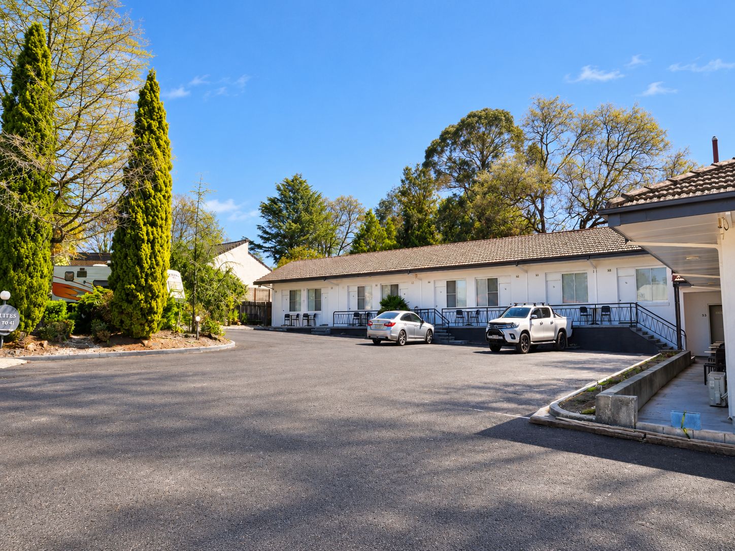 A motel with a parking lot, two cars parked, and a row of rooms with a covered outdoor area.