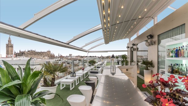 Un bar en la azotea con vistas despejadas a un edificio histórico, asientos blancos y una zona de bar con botellas.