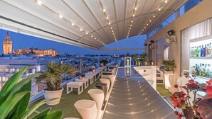 Un bar en la azotea con mesas al aire libre, una zona de bar y vistas a un edificio histórico al fondo.
