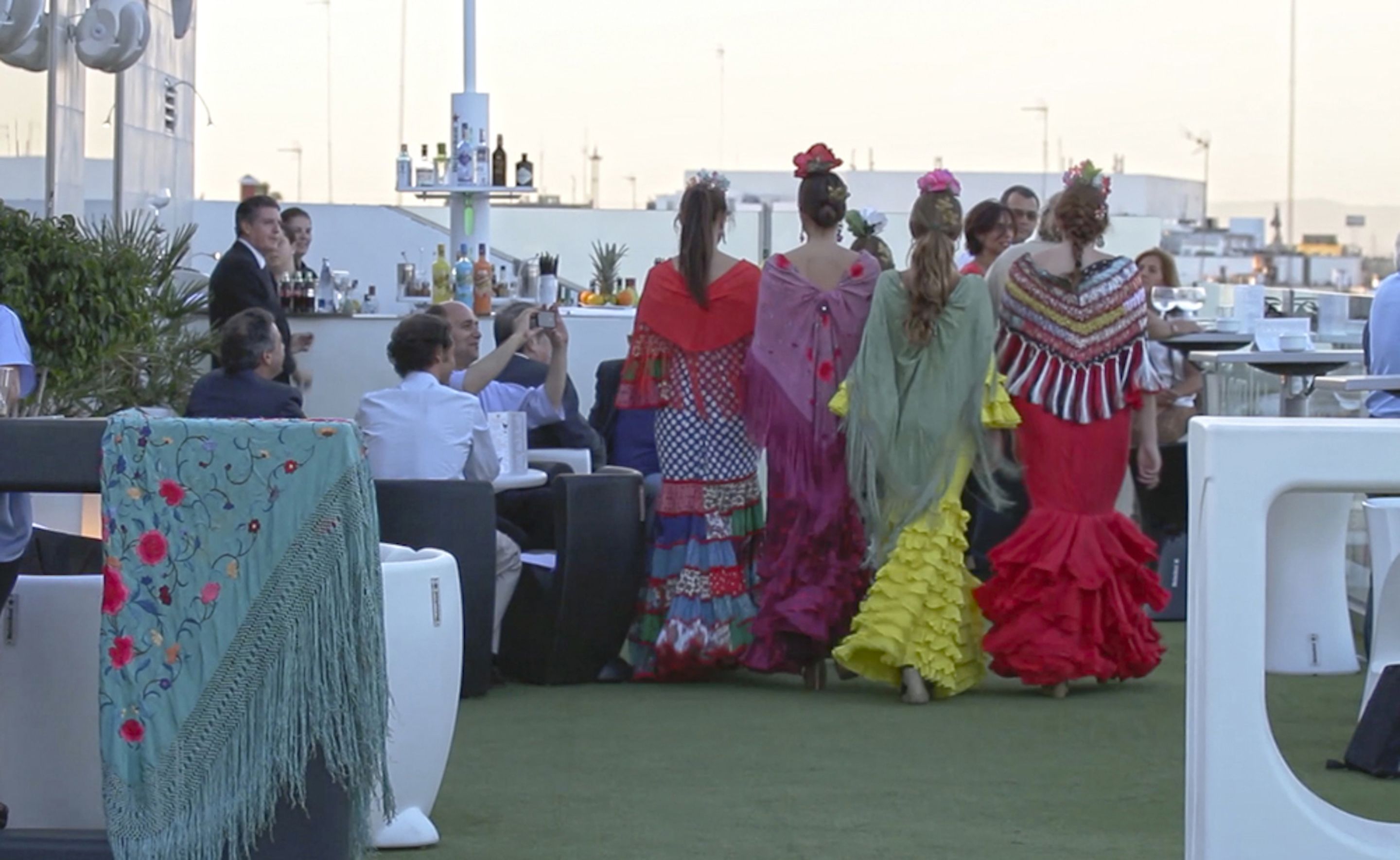 A rooftop event with people in colorful traditional dresses, outdoor seating, and a cityscape in the background.