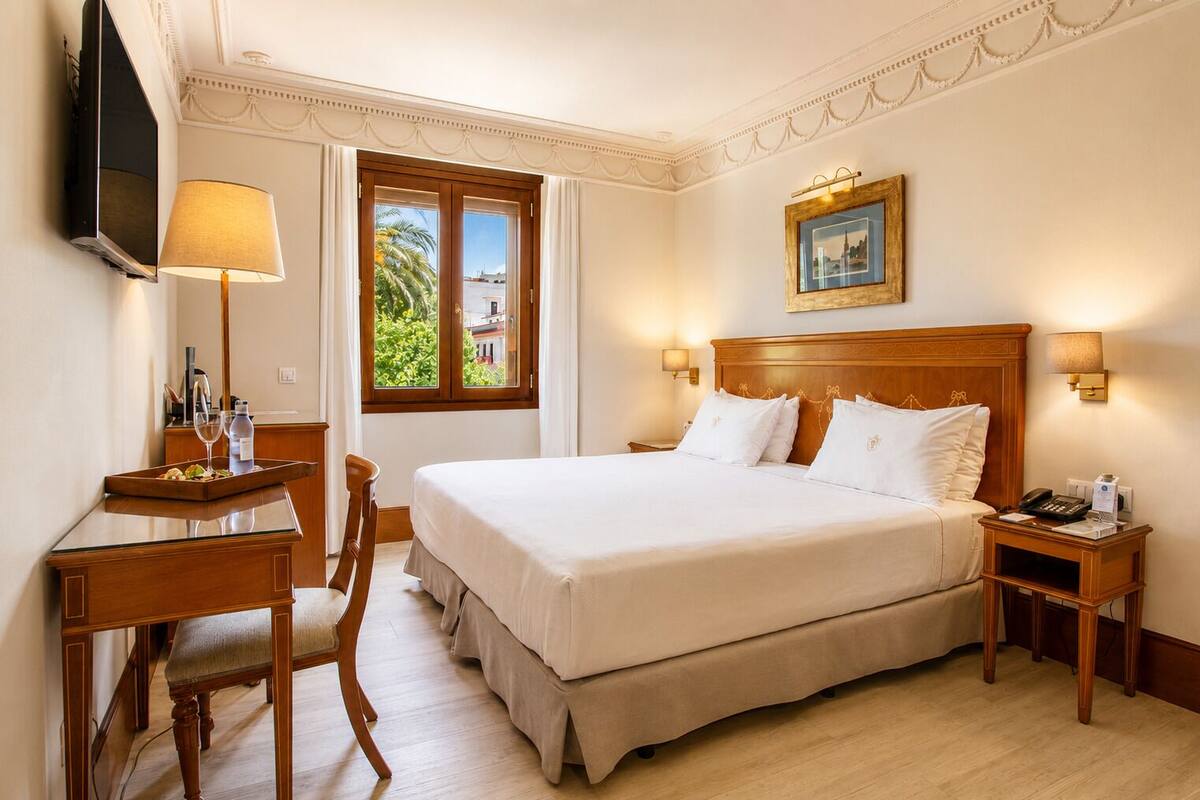 Habitación de hotel con una cama grande, un escritorio con lámpara, una silla, una mesita de noche con teléfono y ventana con vista a zonas verdes.
