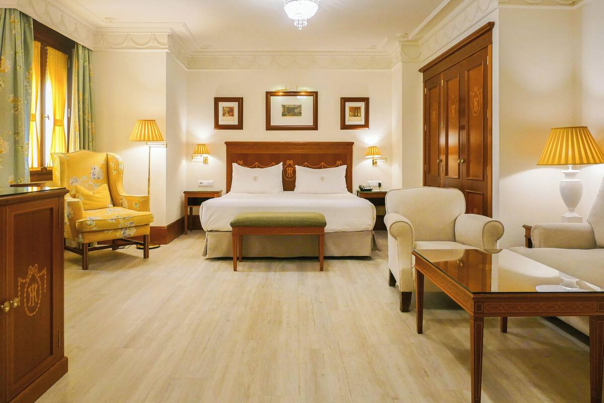 Una habitación de hotel con cama, dos mesitas de noche, un aparador, un sofá, una silla y un armario de madera.