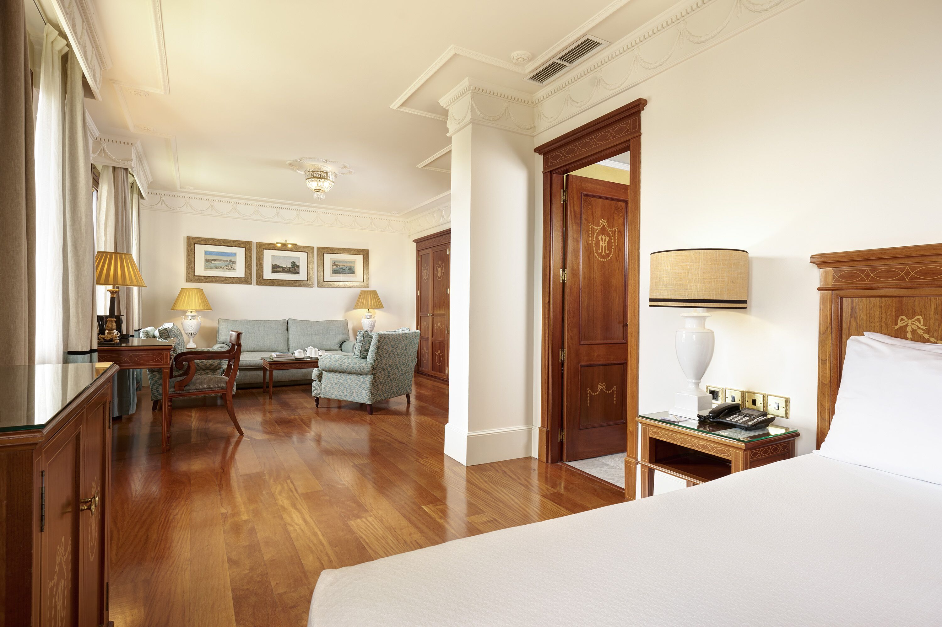 A hotel room with a large bed, a sitting area with a sofa and chairs, a wooden wardrobe, and a desk with a lamp.
