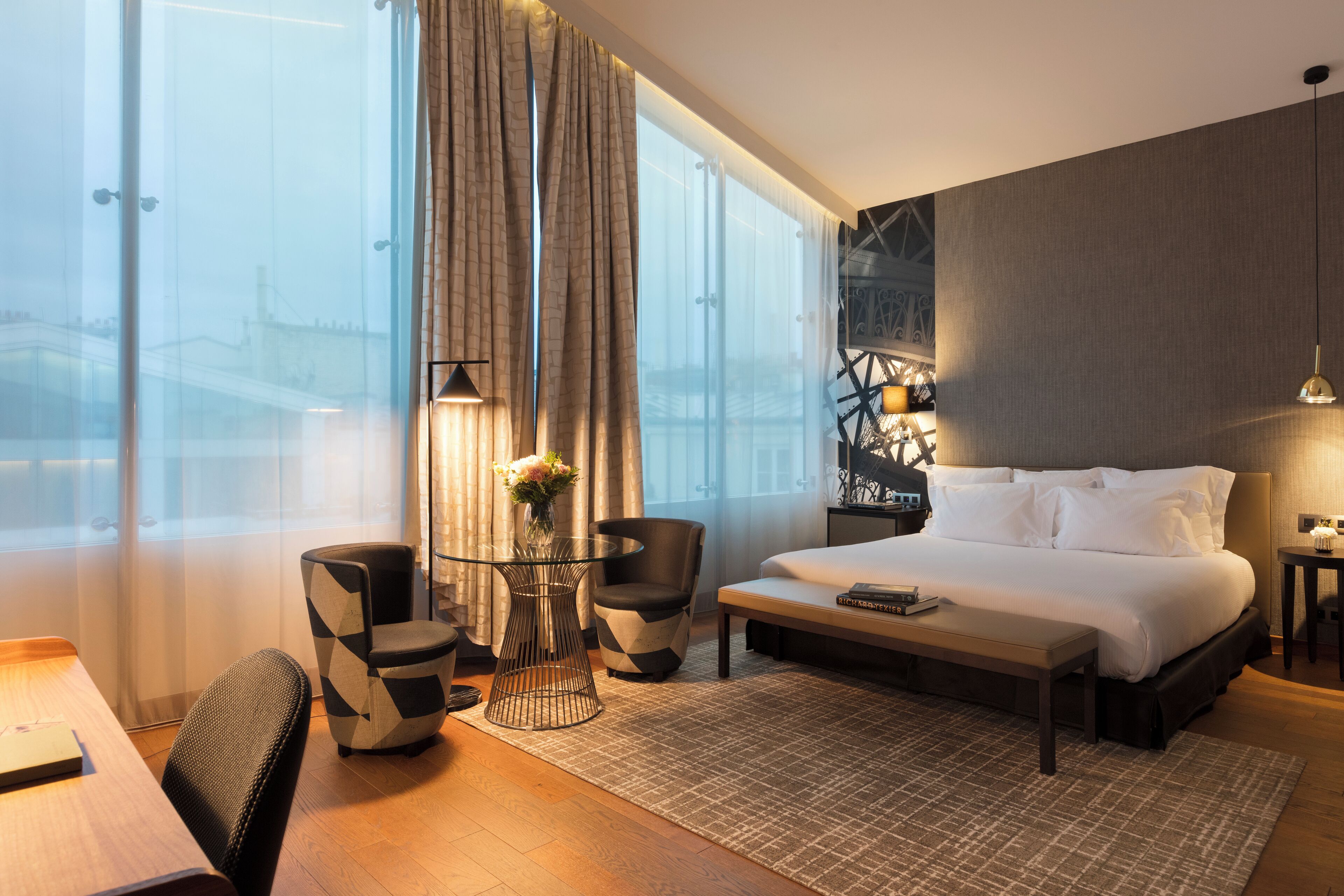 A modern hotel room with a large bed, a desk with a chair, a small table with a vase of flowers, and a view of the city through large windows.