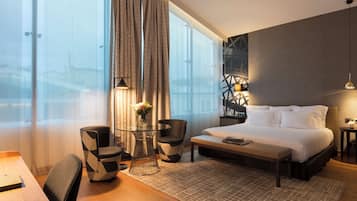 Une chambre d’hôtel moderne dotée d’un grand lit, d’un bureau avec une chaise, d’une petite table ornée d’un vase de fleurs et offrant une vue sur la ville grâce à de grandes fenêtres.