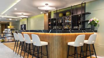 A bar area with a curved counter, white bar stools, and a shelf with bottles and glasses.
