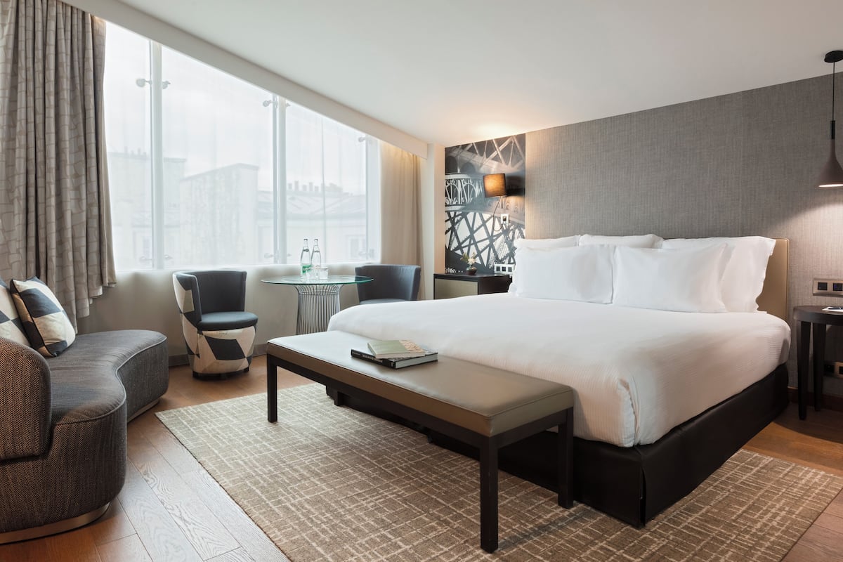 A modern hotel room with a large bed, a seating area with a sofa and chairs, a small table, and a view of a cityscape through large windows.