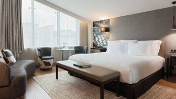 A modern hotel room with a large bed, a seating area with a sofa and chairs, a small table, and a view of a cityscape through large windows.