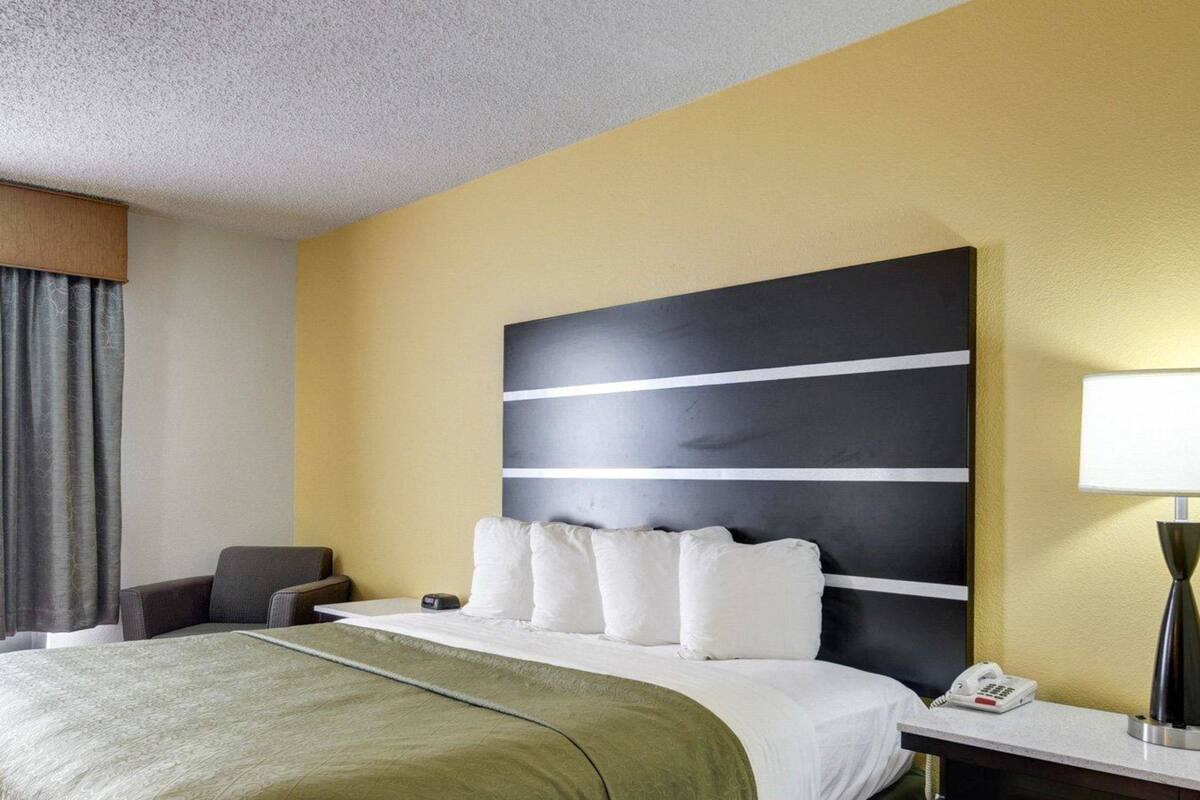 A hotel room with a bed, a bedside table with a lamp, a chair, and a large abstract artwork on the wall.