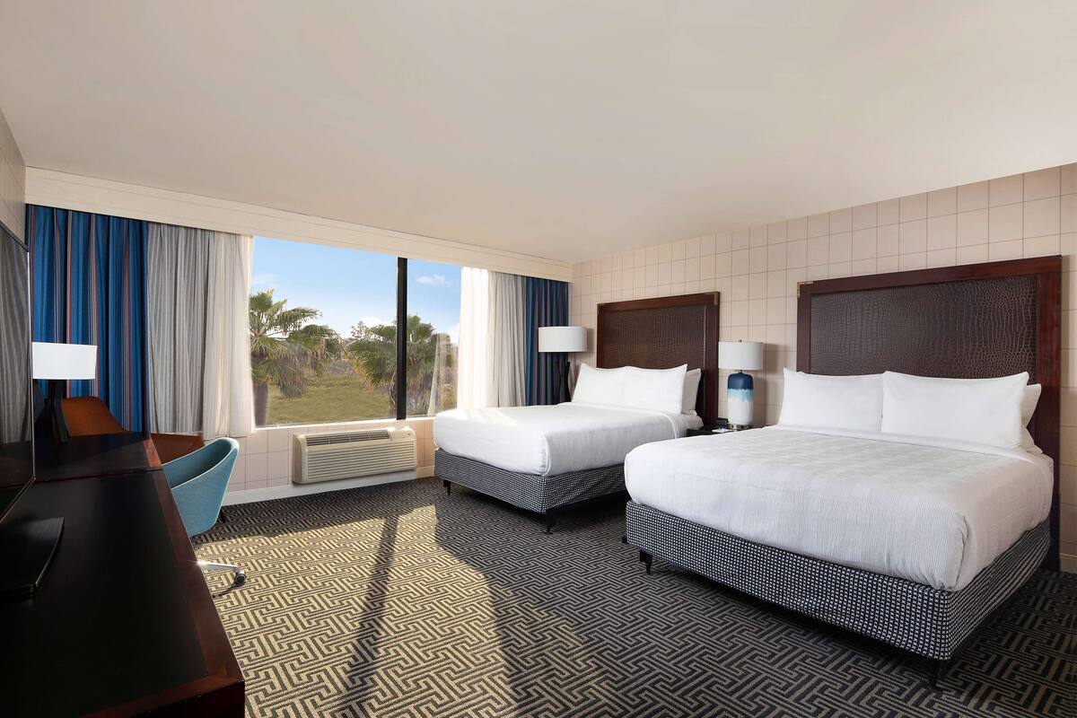 A hotel room with two beds, a desk, and a chair. There are large windows with curtains, a lamp, and a view of palm trees.