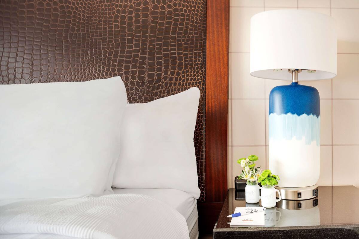 A hotel room with a bed, a nightstand, a lamp, and a small table with a vase of flowers.