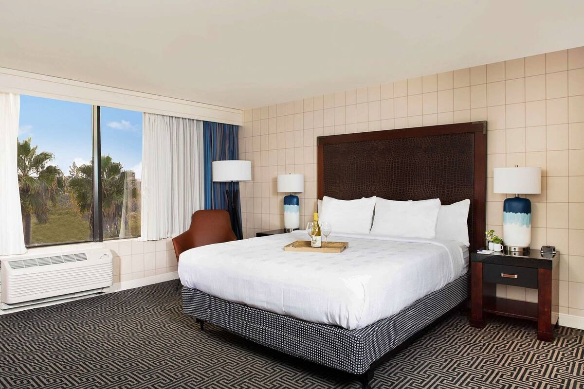 A hotel room with a large bed, two bedside tables, a chair, a window with curtains, and a view of palm trees.