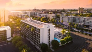 A modern hotel with a rooftop pool, surrounded by palm trees and other buildings.
