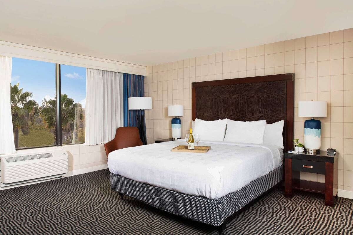 A hotel room with a large bed, two bedside lamps, a nightstand, a chair, and a view of palm trees through the window.