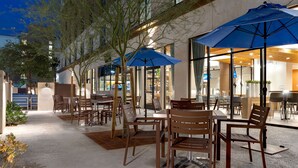 An outdoor seating area with tables and chairs, umbrellas, and a building with large windows in the background. - Home2Suites by Hilton Anaheim Resort (Anaheim)