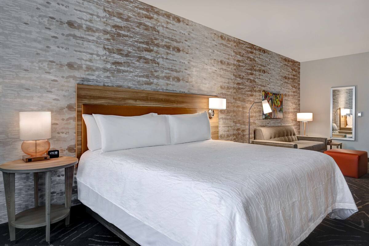 A hotel room with a large bed, two bedside tables with lamps, a small sofa, a wall-mounted artwork, and a mirror.