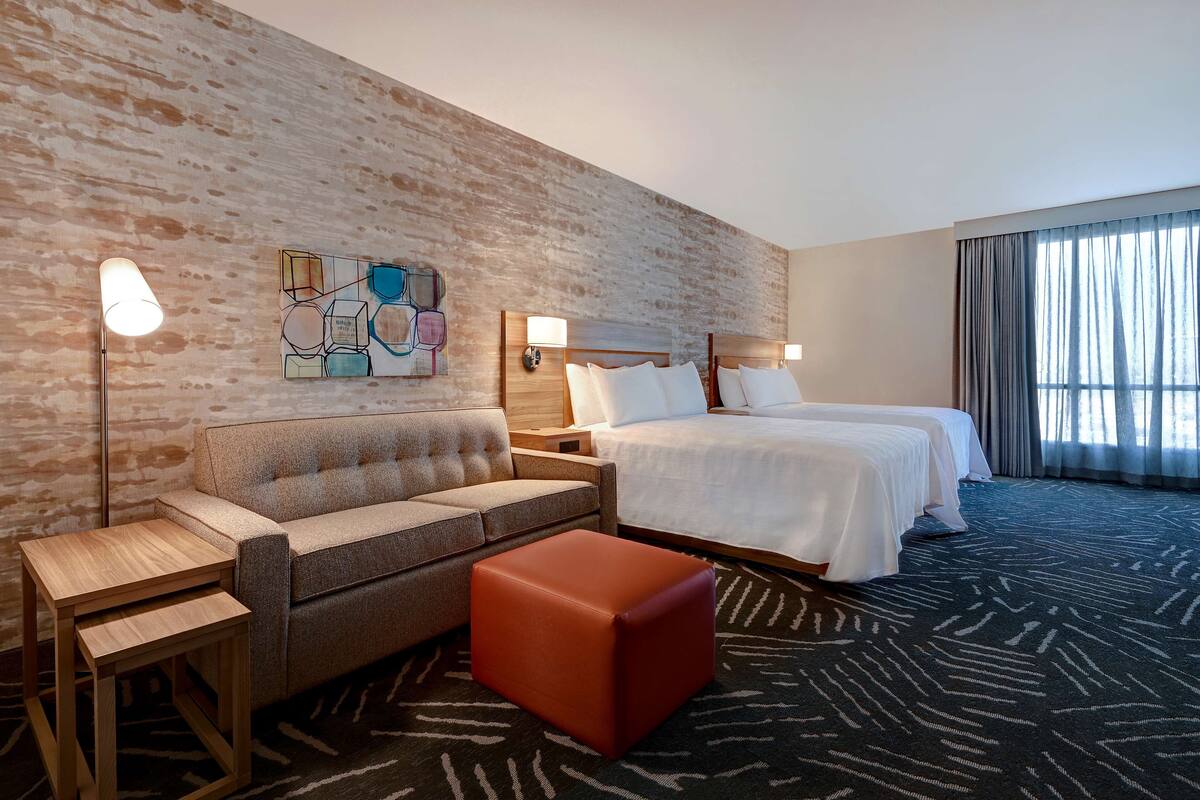 A hotel room with a sofa, a bed, a small table, a red ottoman, and a painting on the wall.