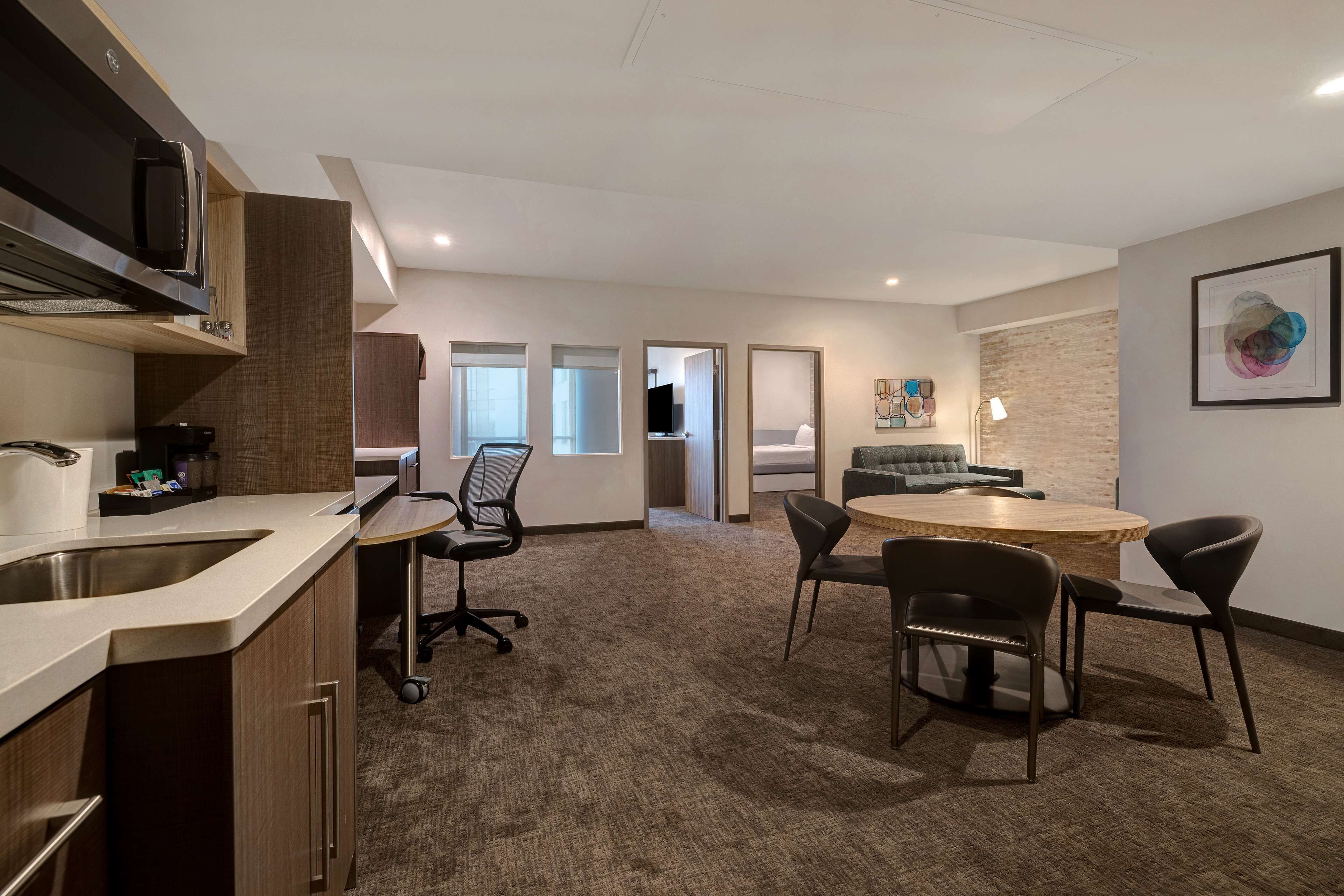 A modern kitchen with a built-in microwave, a sink, and a coffee machine. There is a dining area with a round table and four chairs. The room has a carpeted floor and a large window.