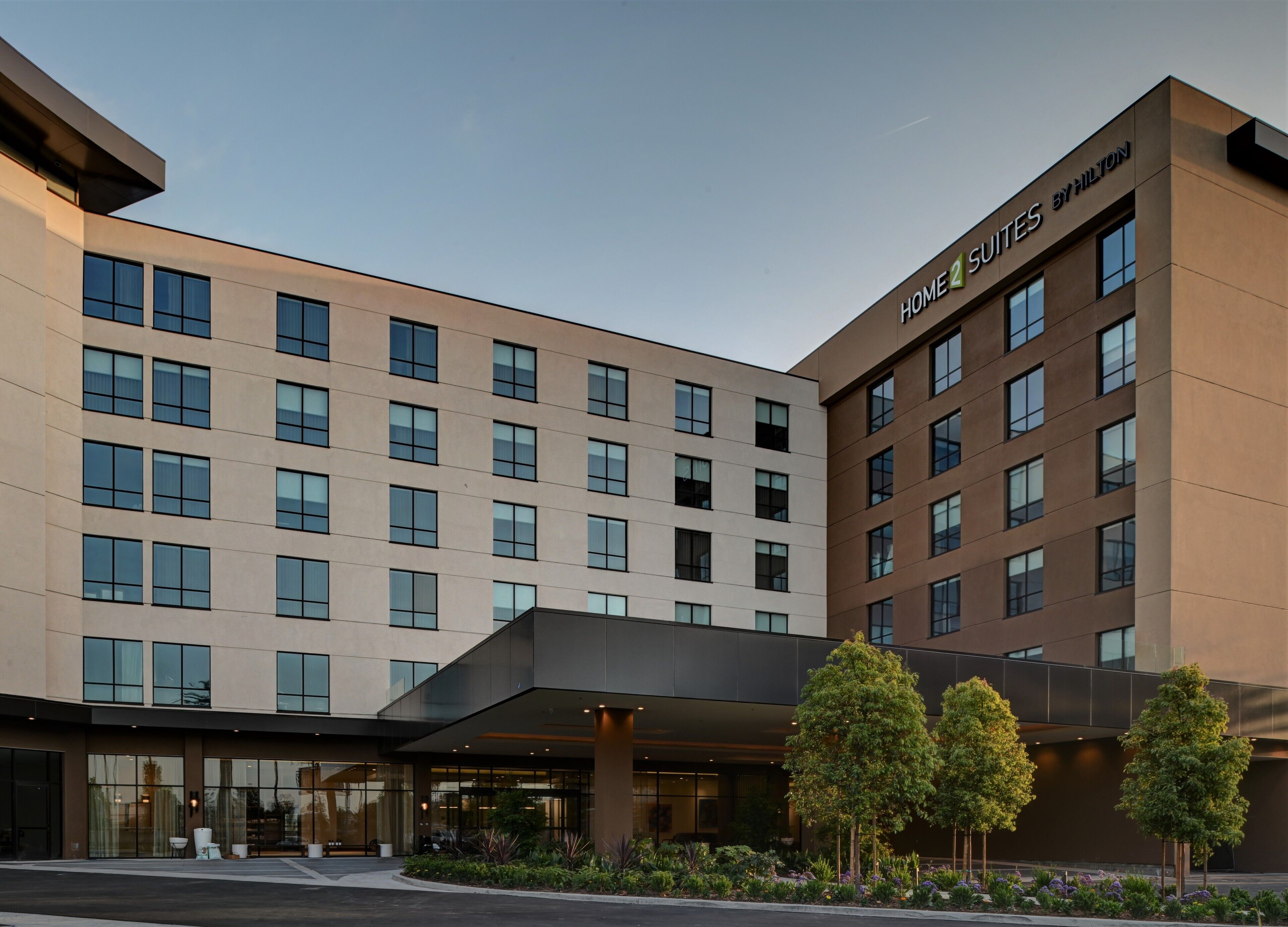 A modern hotel with multiple floors, a prominent entrance with a covered area, and a sign indicating it is a 'Home 2 Suites by Hilton'.