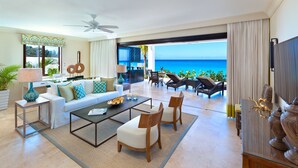A spacious living room with a sofa, coffee table, and chairs, a dining area with a table and chairs, a ceiling fan, and a large window overlooking the ocean. - The Sandpiper (Holetown)