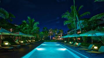 A nighttime pool area with lounge chairs and umbrellas, surrounded by palm trees.