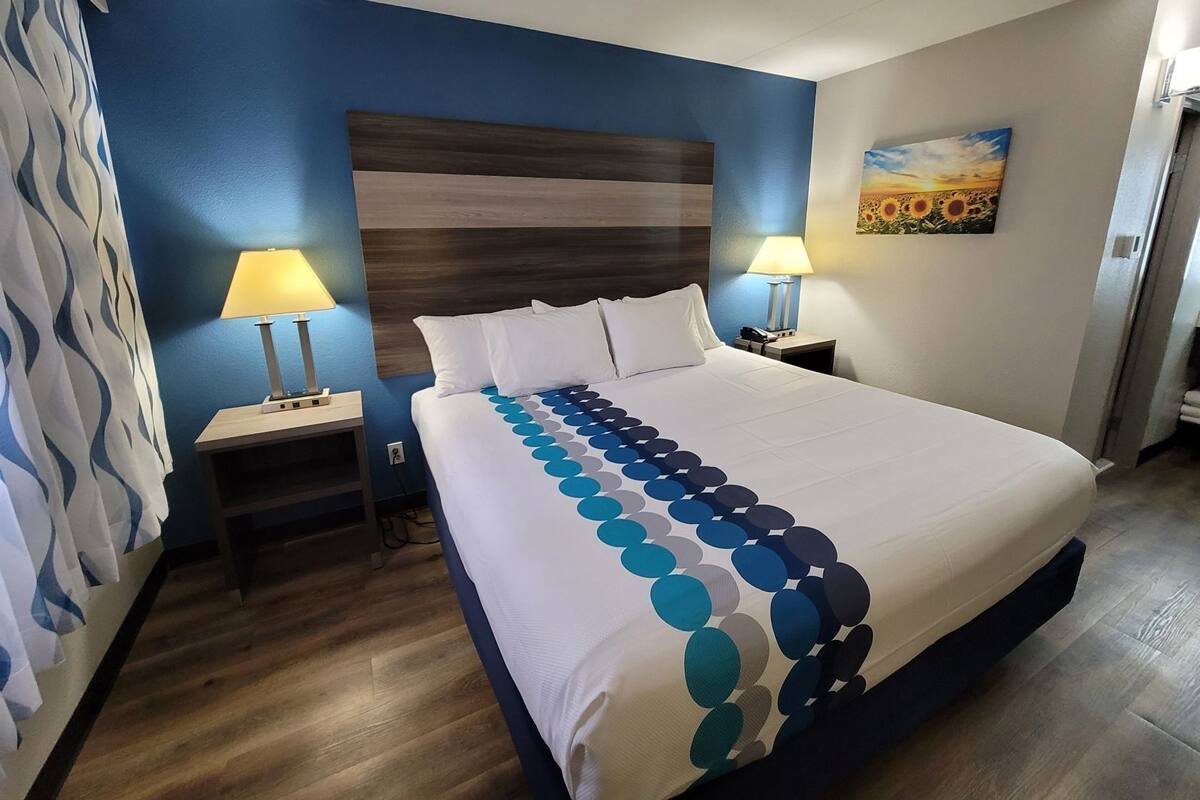A hotel room with a large bed, two bedside lamps, a headboard, a painting on the wall, and a patterned curtain.
