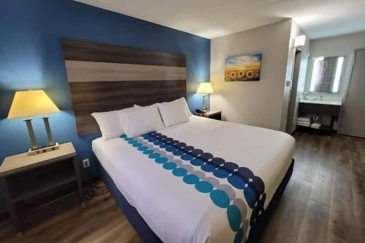 A hotel room with a bed, two bedside tables, a nightstand, a painting on the wall, and a bathroom visible through an open door.