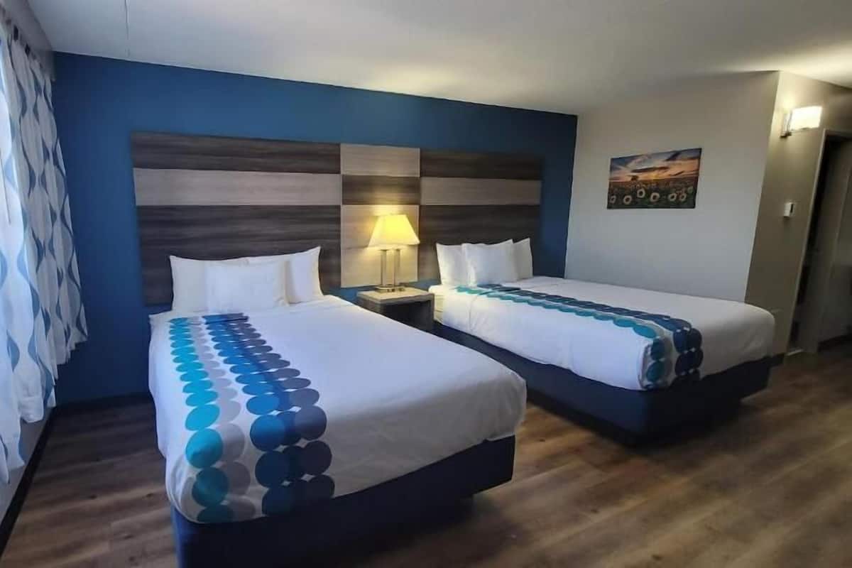 A hotel room with two beds, a wooden floor, a blue accent wall, and a nightstand with a lamp.