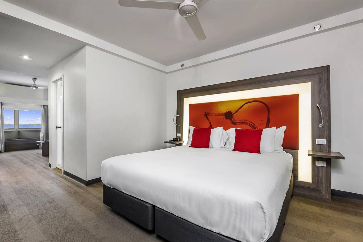Quarto de hotel moderno com cama grande, ventilador de teto, vista para o mar e cabeceira de madeira.