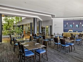 Un comedor moderno con mesas y sillas, una cocina al fondo y vistas al exterior con vegetación.