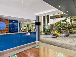A modern hotel reception area with a blue counter, reception desk, and seating area with plants.