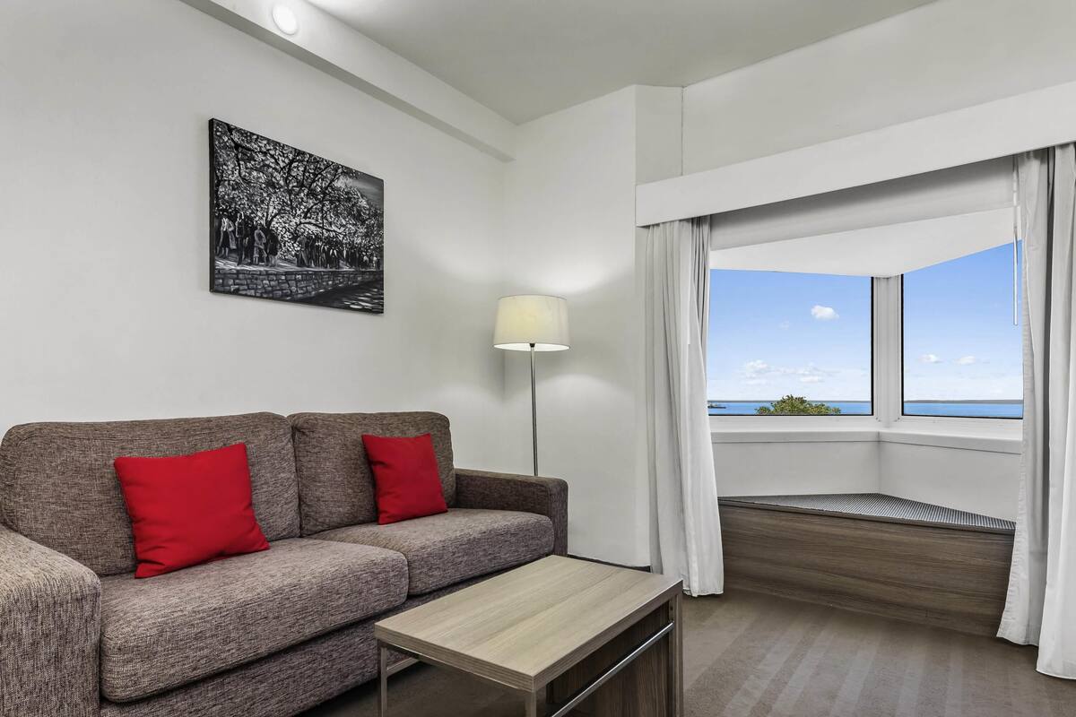 Quarto de hotel moderno com sofá cinza, uma pequena mesa de madeira, luminária de chão, uma janela grande com cortinas e uma obra de arte em preto e branco na parede.