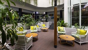 A spacious indoor area with multiple seating arrangements, including white wicker chairs and round tables, surrounded by lush green plants. - Novotel Darwin CBD (Darwin)