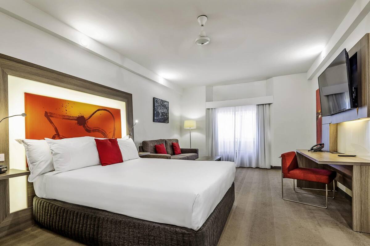 Quarto de hotel com uma cama grande, uma escrivaninha com cadeira, televisão e janela com cortinas.