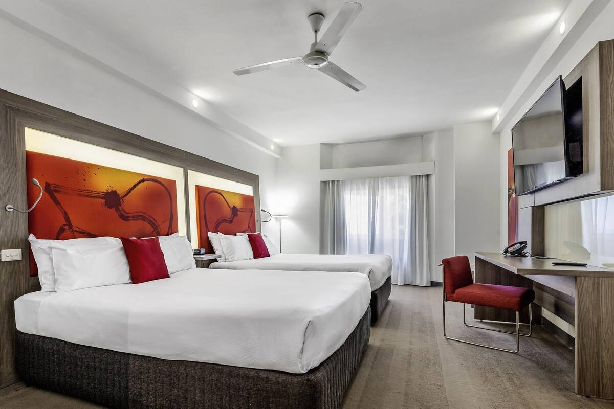 Quarto de hotel com duas camas, uma escrivaninha, uma cadeira, ventoinha no teto e obras de arte nas paredes.