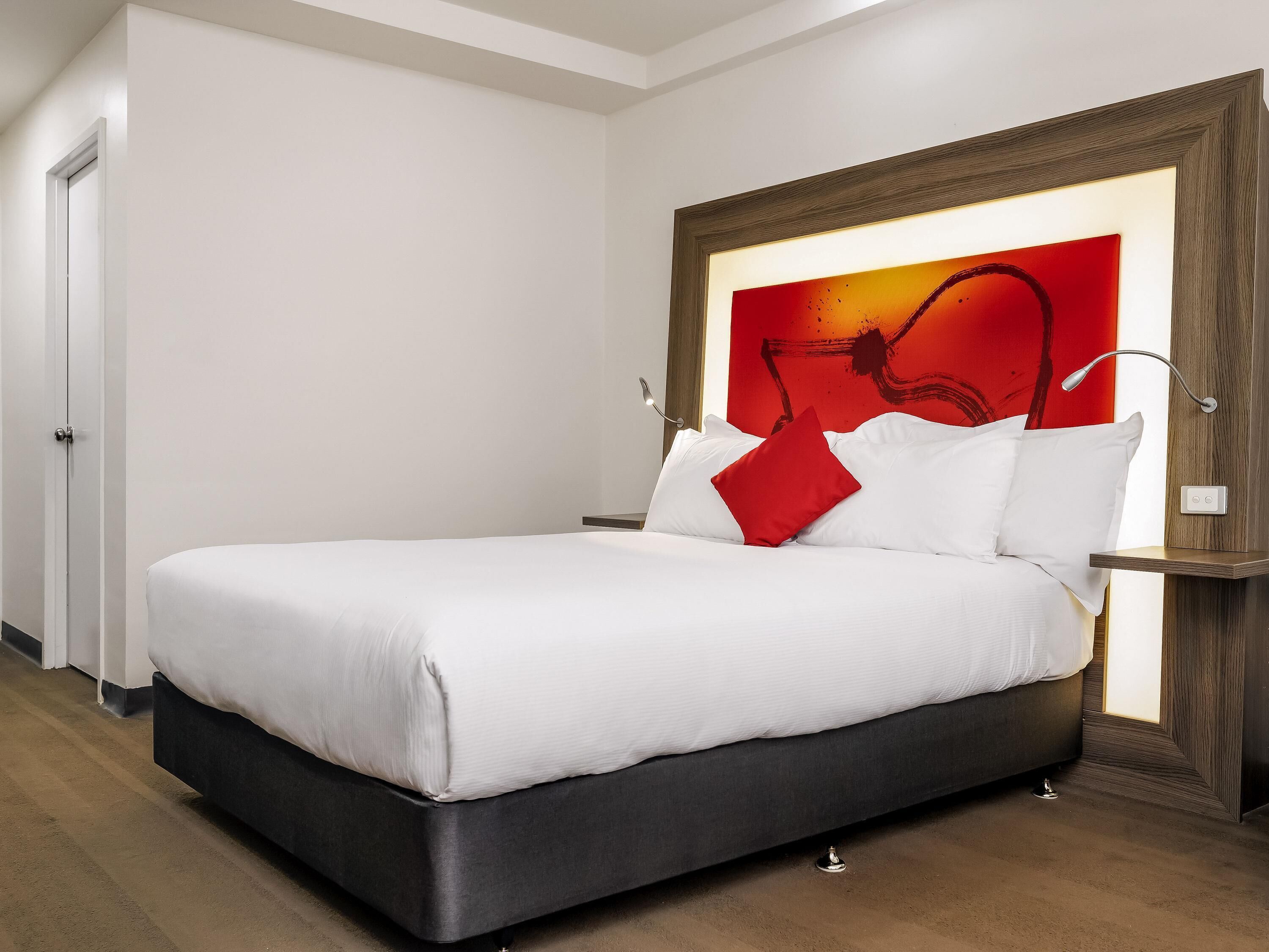A modern hotel room with a large bed, a wooden headboard, and a red abstract painting on the wall.