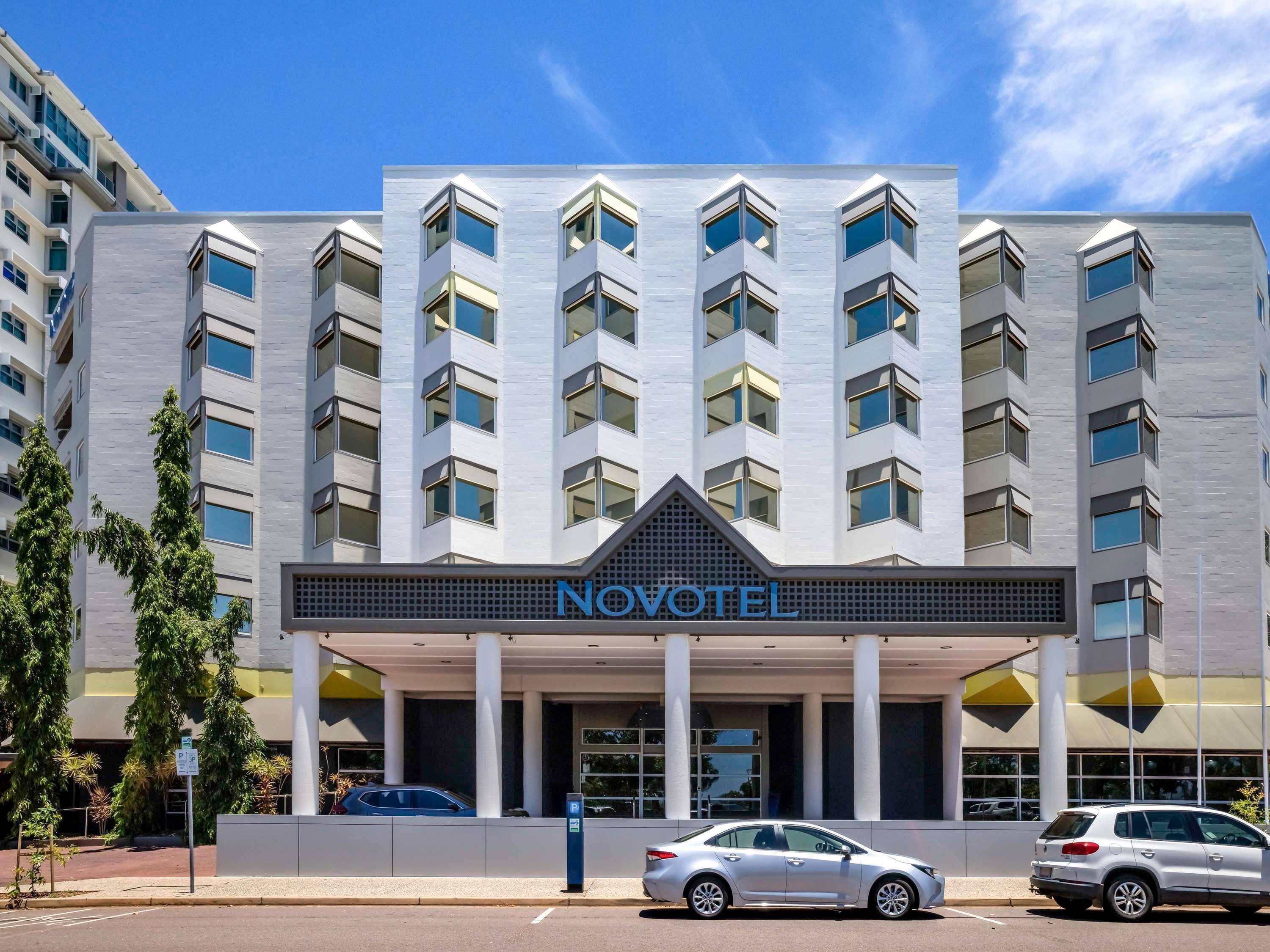 A modern hotel building with a prominent 'NOVOTEL' sign, multiple floors, and a parking area with cars.