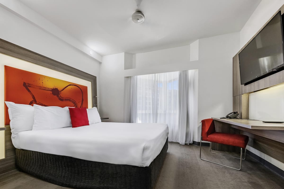 Quarto de hotel com cama, escrivaninha, cadeira, televisão e uma janela grande com cortinas.