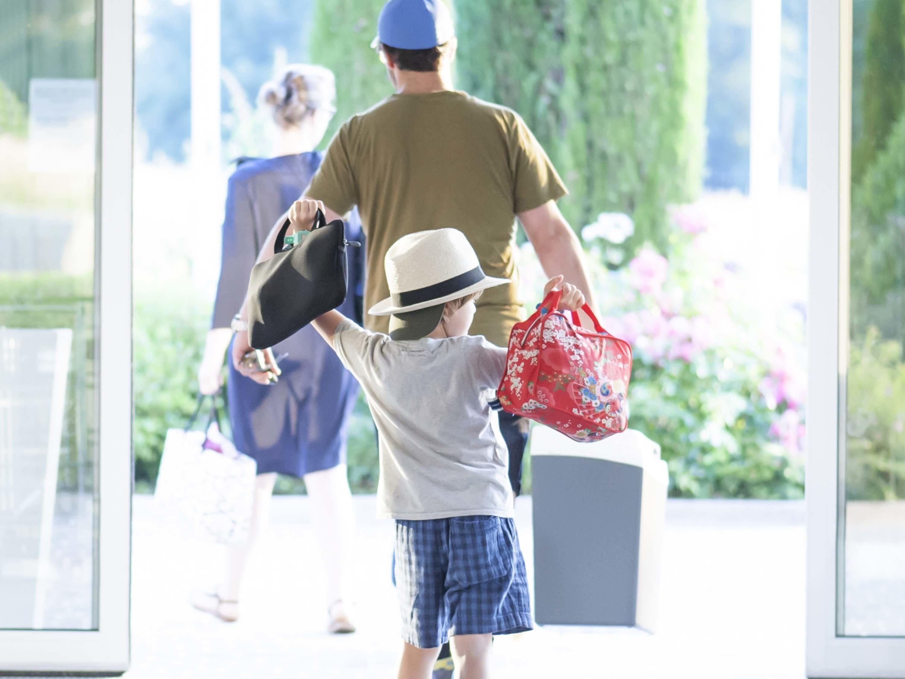 Un enfant tenant un sac rouge et un chapeau, avec un adulte en arrière-plan.