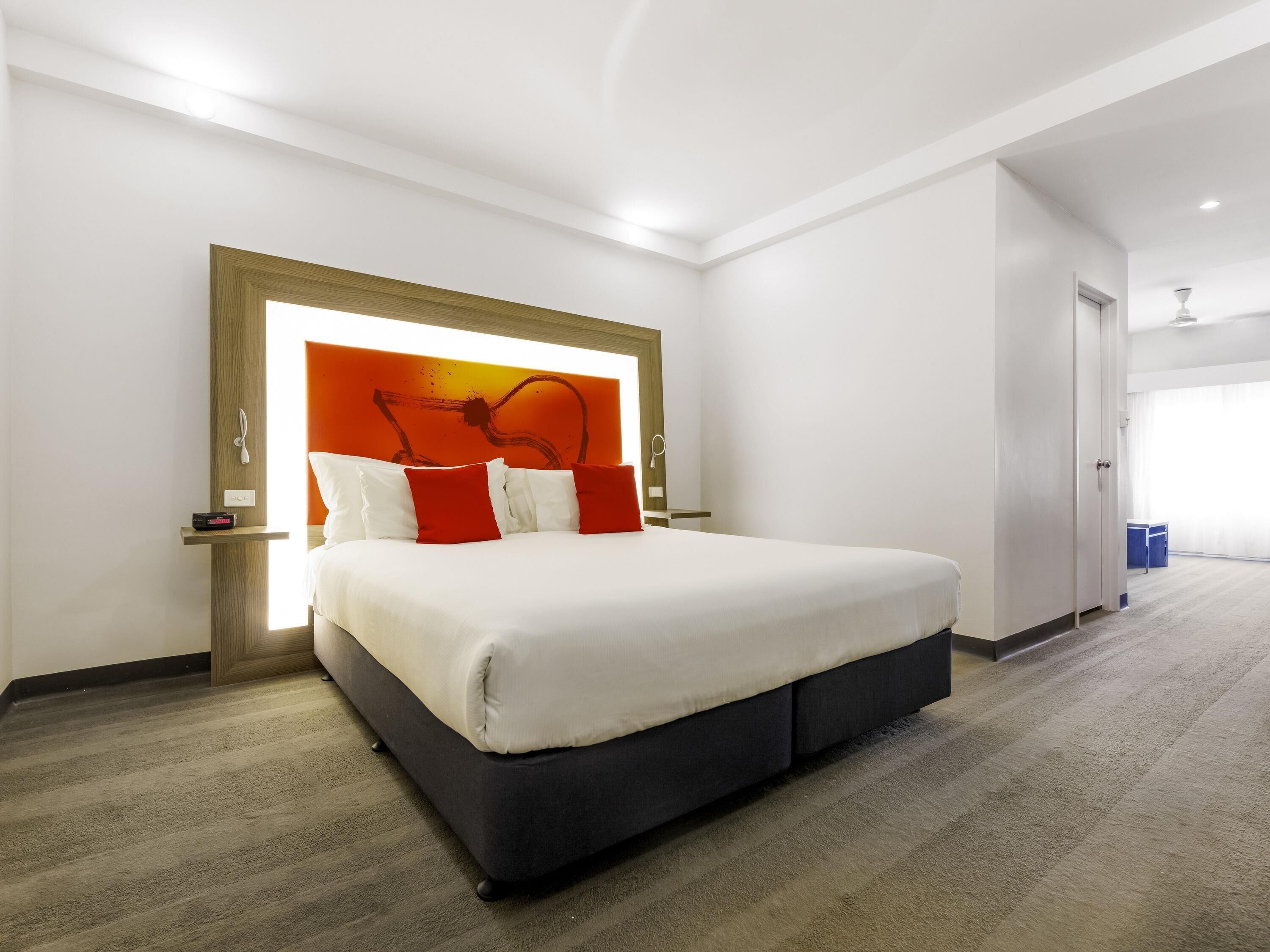 Una habitación de hotel moderna con una cama grande, un cabecero con un diseño de iluminación único, una mesita de noche y suelo alfombrado.