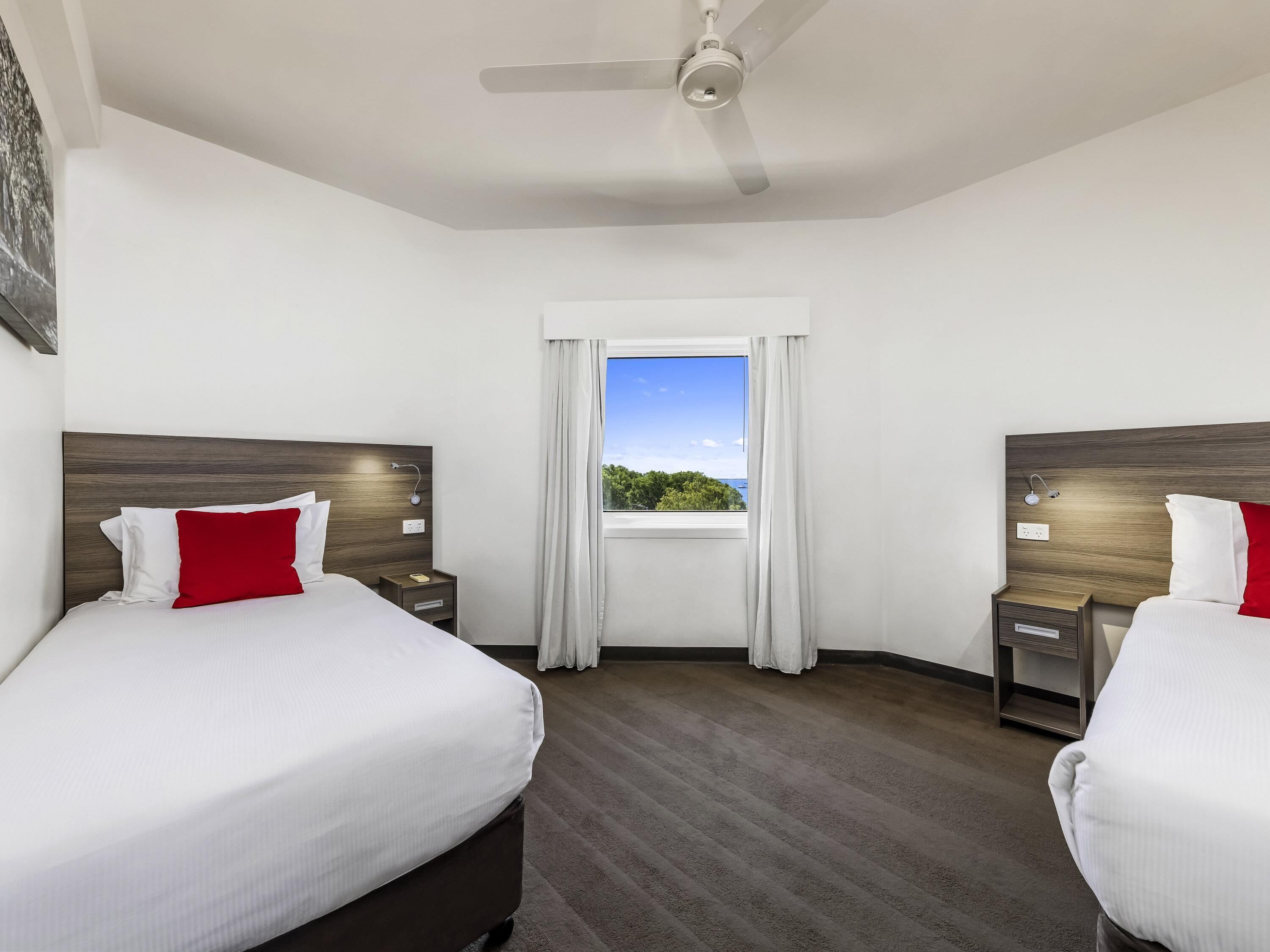 A hotel room with two beds, a ceiling fan, a window with a view, and a nightstand with a lamp.