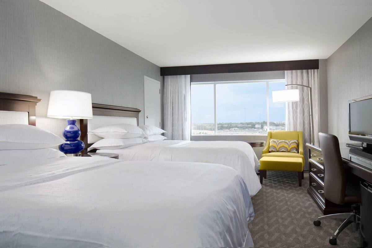 A hotel room with two beds, a desk, a chair, and a large window offering a city view.