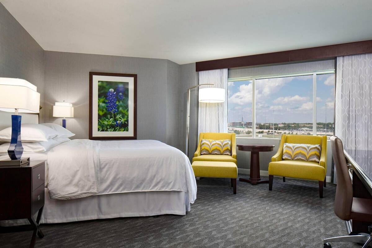 A hotel room with a large bed, two yellow armchairs, a small table, and a view of the city through large windows.