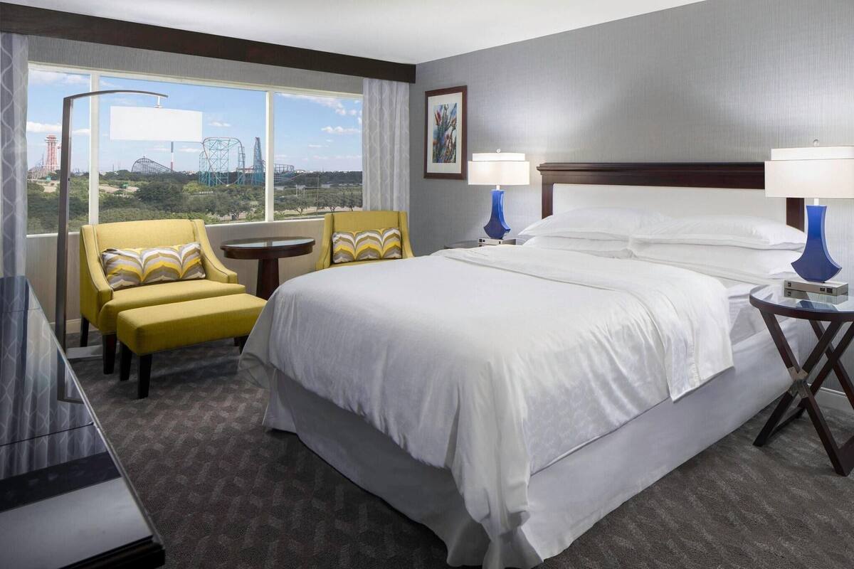 A hotel room with a large bed, two bedside tables with lamps, a yellow armchair, a bench, and a view of a theme park.