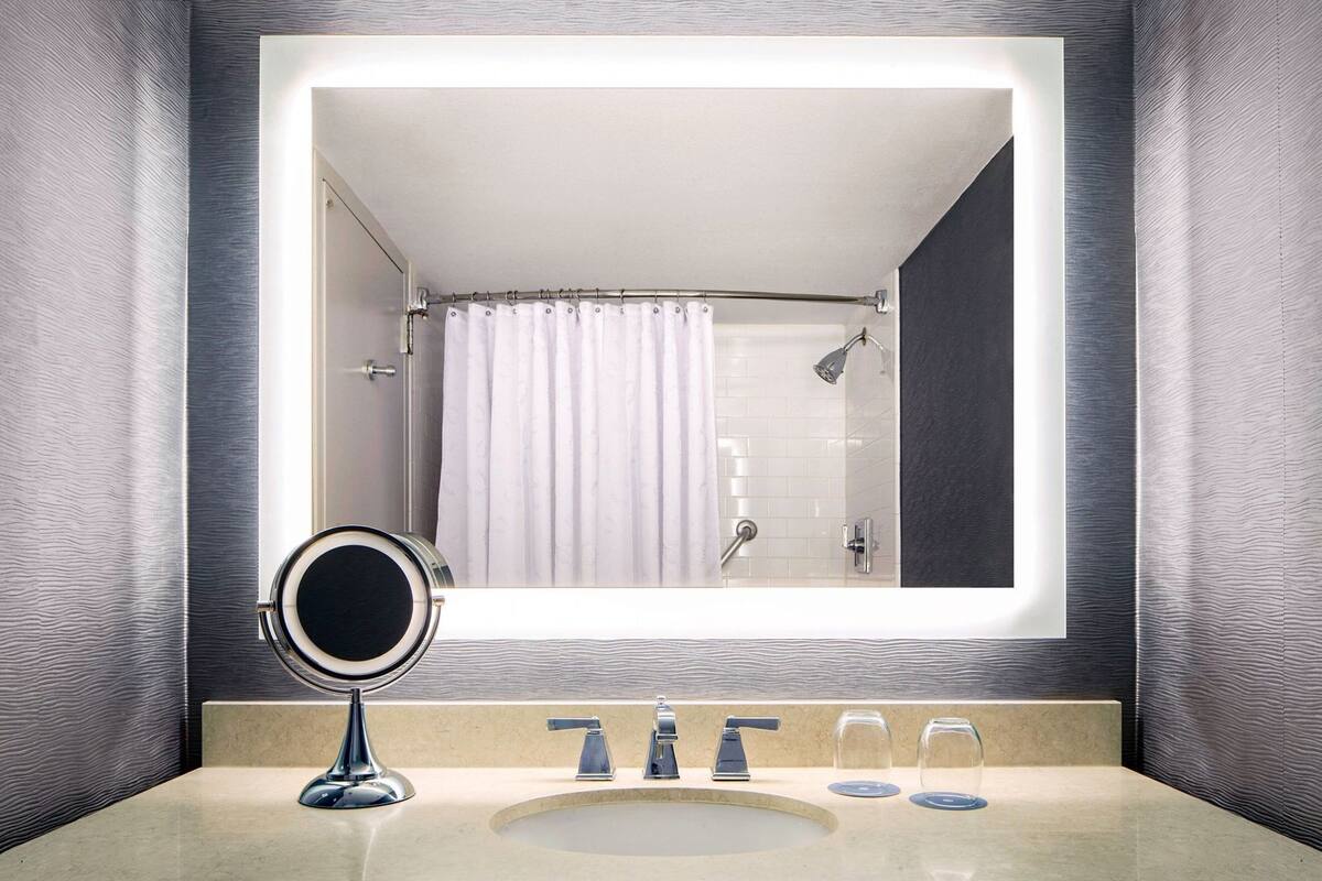 A bathroom with a large mirror, a sink with two faucets, and a shower area.