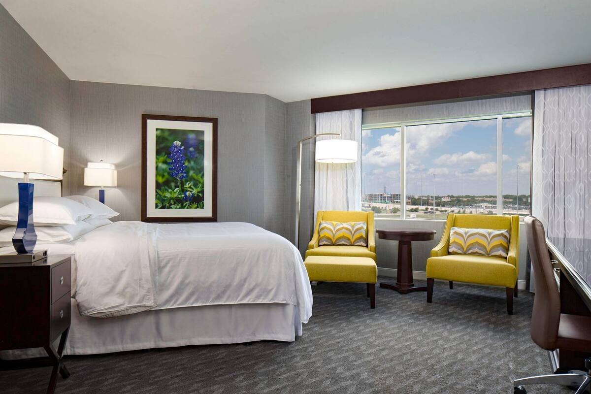 A hotel room with a bed, two yellow armchairs, a small table, a lamp, a framed picture, and a large window with a view.