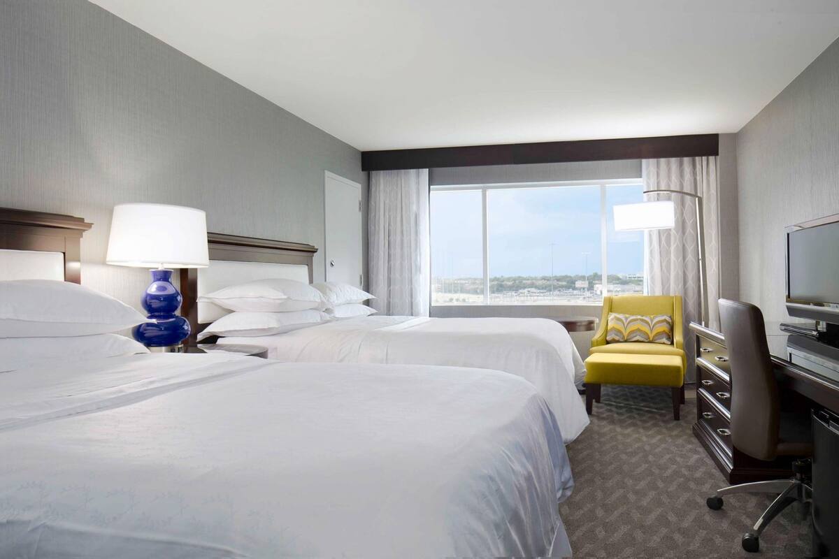 A hotel room with two beds, a desk with a TV, a chair, and a large window with a city view.